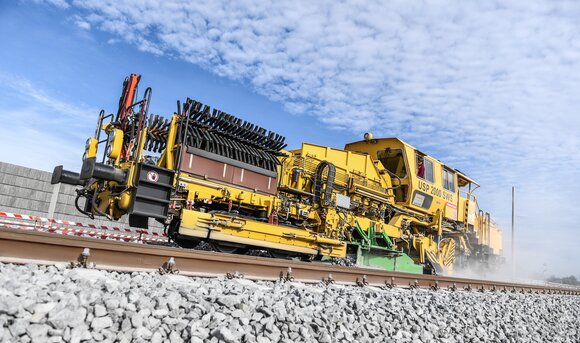 Ein Baustellenfahrzeug der Deutschen Bahn fährt vor blauem Himmel über die Schiene. 