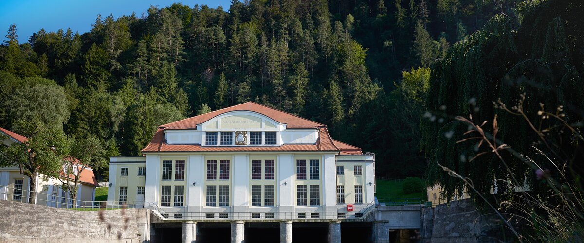 Ein Haus ist an einem Fluss gelegen, dahinter erstreckt sich ein Wald aus Nadelbäumen. | © DB AG / Faruk Hosseini