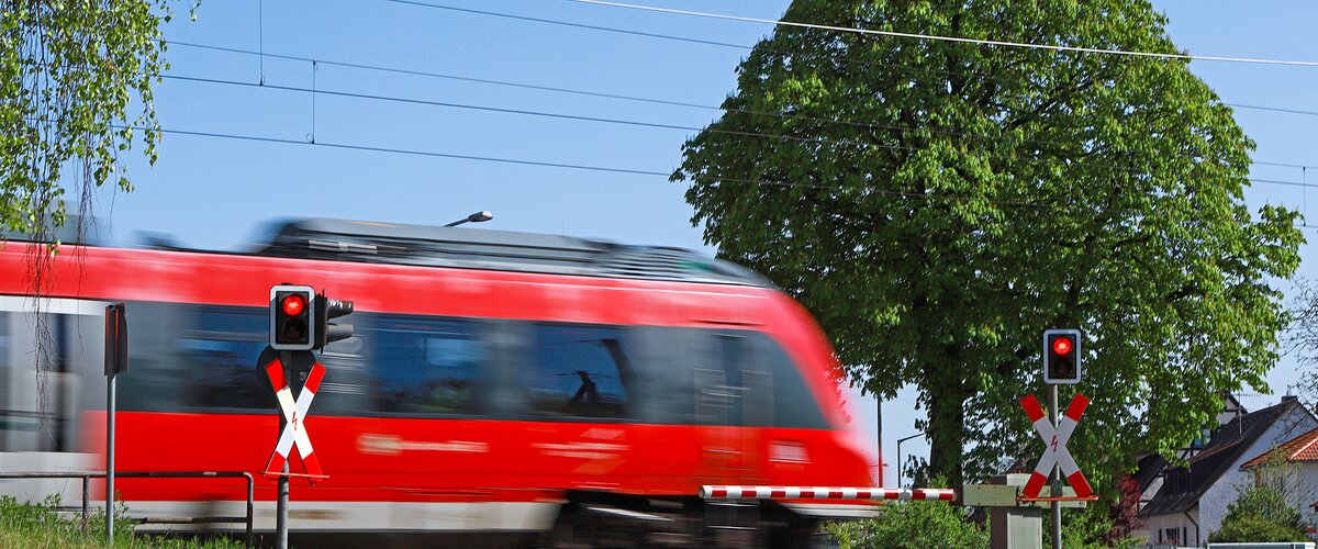 A red train crosses a level crossing in summer, the barriers are closed. | © Deutsche Bahn AG / Claus Weber 