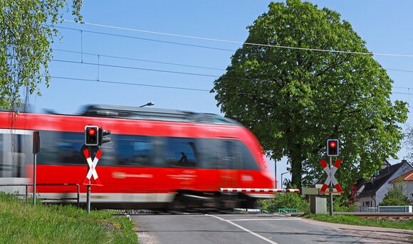 Ein roter Zug fährt im Sommer über einen Bahnübergang, die Schranken sind geschlossen. 