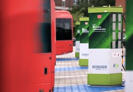 Eine grüne Ladesäule neben roten Elektro-Bussen am Bahnhof. 