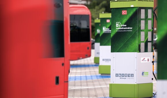 Eine grüne Ladesäule neben roten Elektro-Bussen am Bahnhof. 