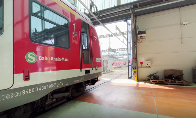 Ein roter Zug steht auf Gleisen in in der Halle eines Instandhaltungswerks, die Hallentore schließen sich hinter ihm. 