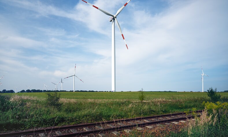 Windräder stehen auf einem Feld neben einer Gleisanlage.