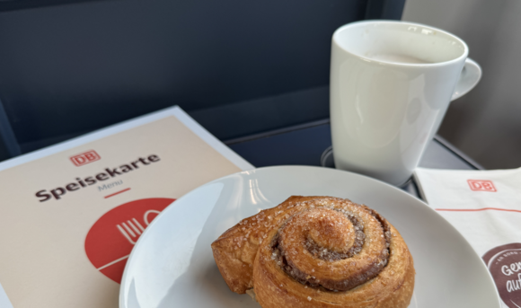 Ausgeklappter Tisch im Zug mit einem Teller mit Zimtschnecke und einer Serviette und Speisekarte des Bordbistros