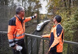 Zwei Mitarbeiter betreiben an einem Gleis Vegetationspflege, während ein Zug vorbei fährt.