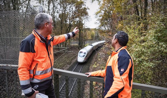 Zwei Mitarbeiter betreiben an einem Gleis Vegetationspflege, während ein Zug vorbei fährt.