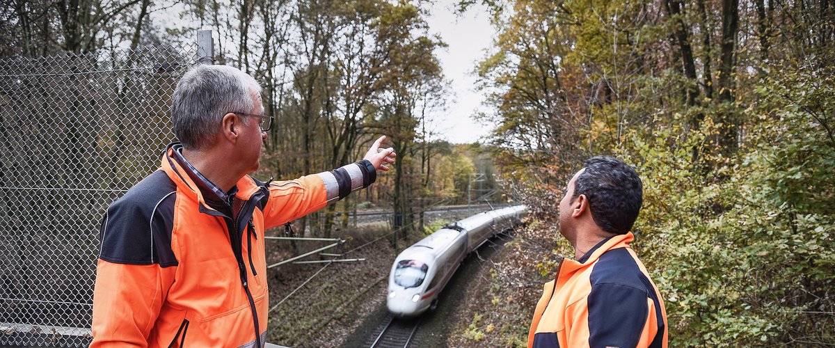 Zwei Mitarbeiter betreiben an einem Gleis Vegetationspflege, während ein Zug vorbei fährt. | © DB AG / Thomas Herter
