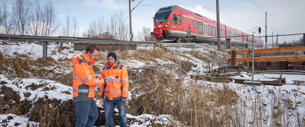 Zwei DB-Mitarbeiter beraten sich an einer Baustelle an einer Bahnstrecke, während ein Zug vorbeifährt. | © DB AG / Faruk Hosseini