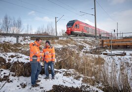 Zwei DB-Mitarbeiter beraten sich an einer Baustelle an einer Bahnstrecke, während ein Zug vorbeifährt.