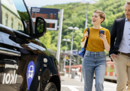 Eine Frau und ein Mann laufen an einem Auto vorbei und schauen auf das Smartphone.