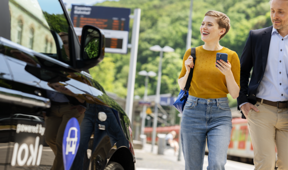 A woman and a man walk past a car and look at their smartphone.