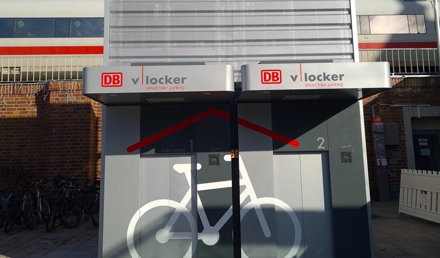 Zwei automatisierte Fahrradparkhäuser für insgesamt zwölf Räder stehen vor dem Zukunftsbahnhof in Halle (Saale). Im Hintergrund fährt ein Zug vorbei.