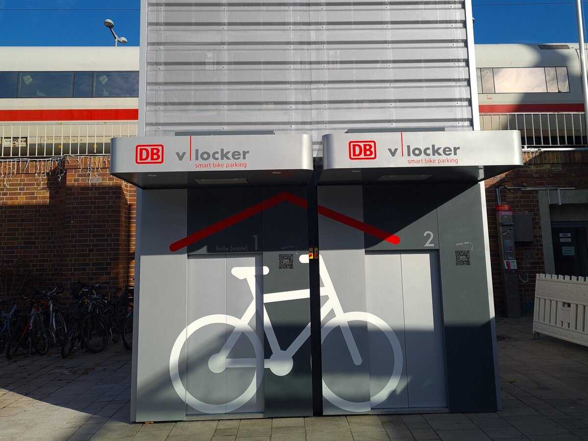 Zwei automatisierte Fahrradparkhäuser für insgesamt zwölf Räder stehen vor dem Zukunftsbahnhof in Halle (Saale). Im Hintergrund fährt ein Zug vorbei. | © DB AG