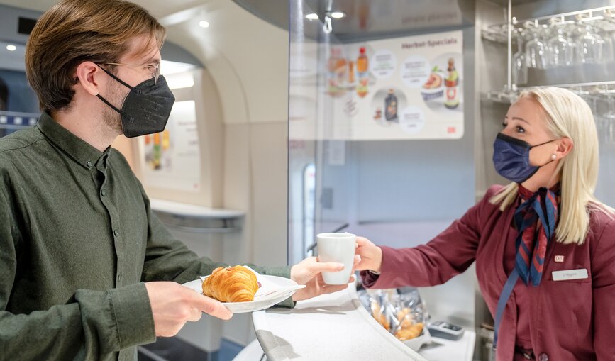 Eine Mitarbeiterin des Bordbistros serviert einem Bahnkunden seine Bestellung auf Porzellangeschirr