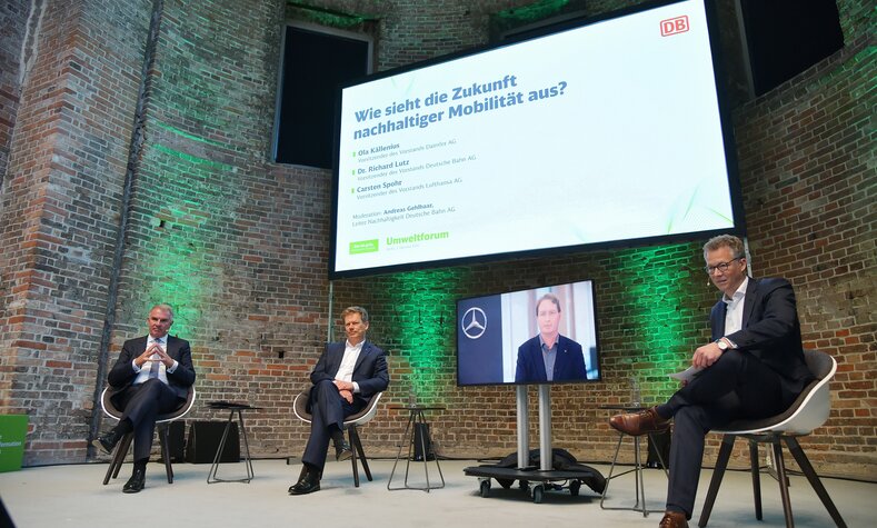 Drei Männer sitzen in einem Gesprächspanel auf einer Bühne. Ein weiterer Mann ist virtuell per Leinwand dazugeschaltet. 