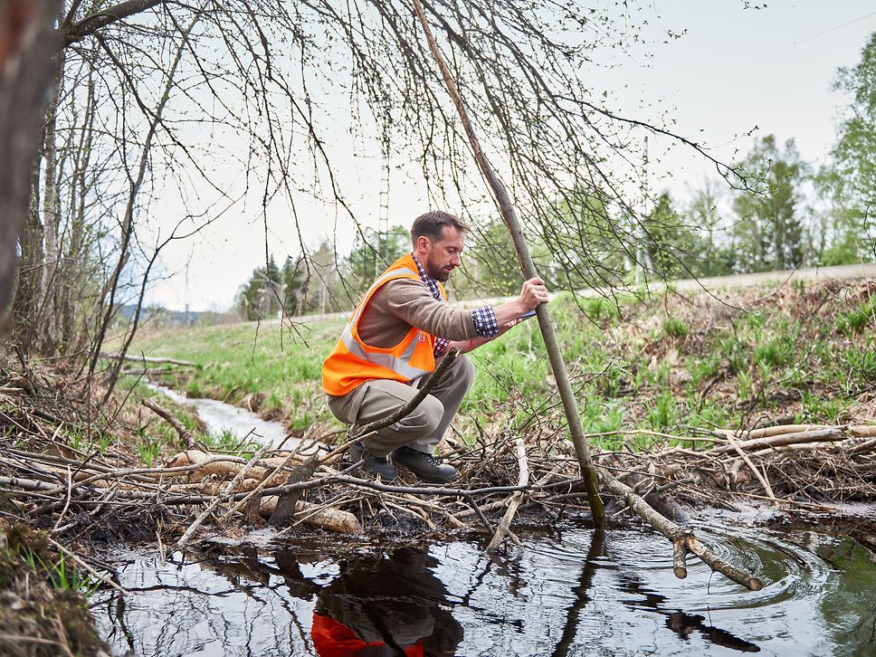 Eine Person in Warnweste hockt an einem Biberdamm und hält einen langen Stock ins Wasser.