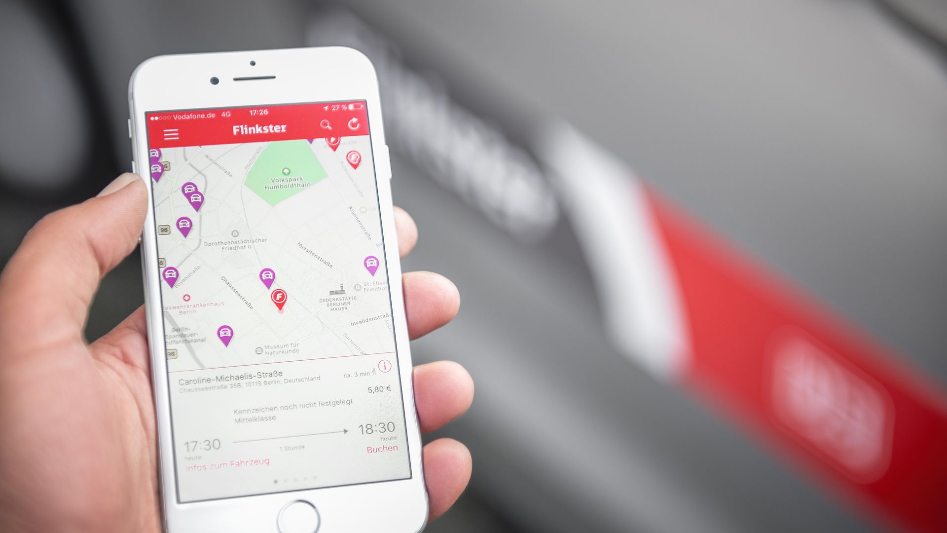 Eine Hand hält ein Smartphone mit geöffneter Flinkster-App. Im Hintergrund ist unscharf ein Fahrzeug mit DB-Logo zu sehen.