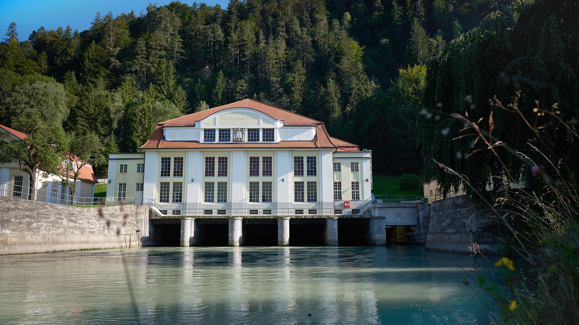 Historisches Saalachkraftwerk in Bad Reichenhall mit hellem Gebäude am Flussufer, umgeben von bewaldeten Hügeln.