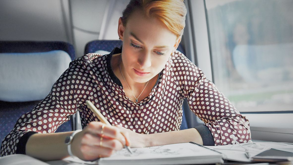 Eine Geschäftsreisende sitzt im ICE am Fenster und schreibt etwas in ein Notizbuch.