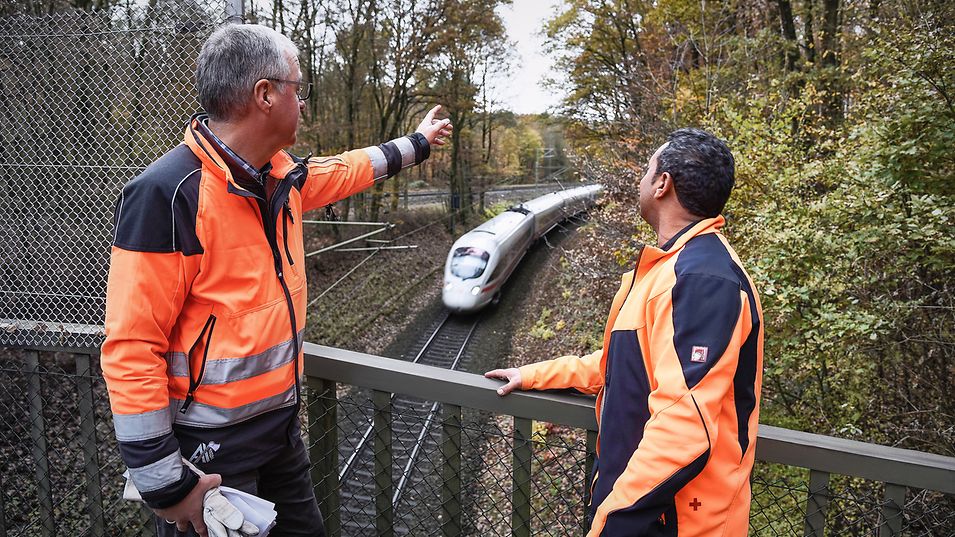 Zwei Bahnmitarbeiter stehen auf einer Brücke und schauen auf einen vorbeifahrenden ICE