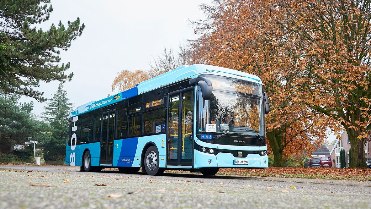 Ein hellblauer Stadtbus steht an einer Straße mit herbstlichen Bäumen im Hintergrund