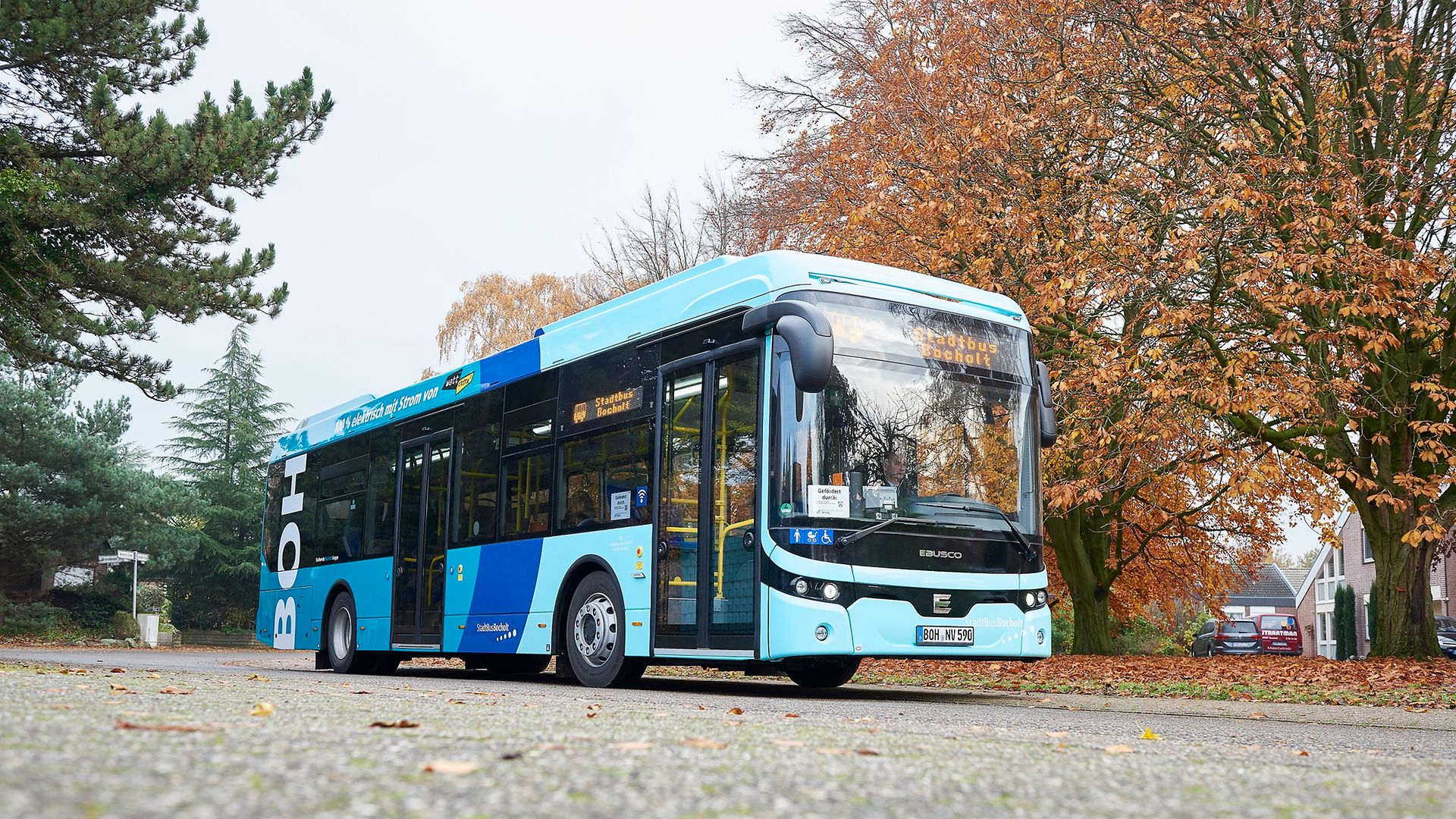 Ein hellblauer Stadtbus steht an einer Straße mit herbstlichen Bäumen im Hintergrund
