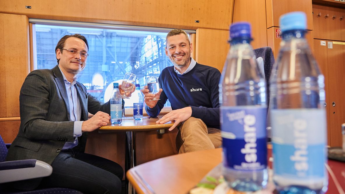 Zwei Männer sitzen im Borbistro und stoßen mit Wasser aus den share -Flaschen an