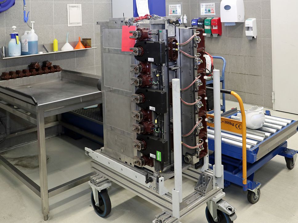 Großes elektrisches Bauteil auf einem Rollwagen in einer Werkstatt, umgeben von Arbeitsmitteln und Reinigungsutensilien.