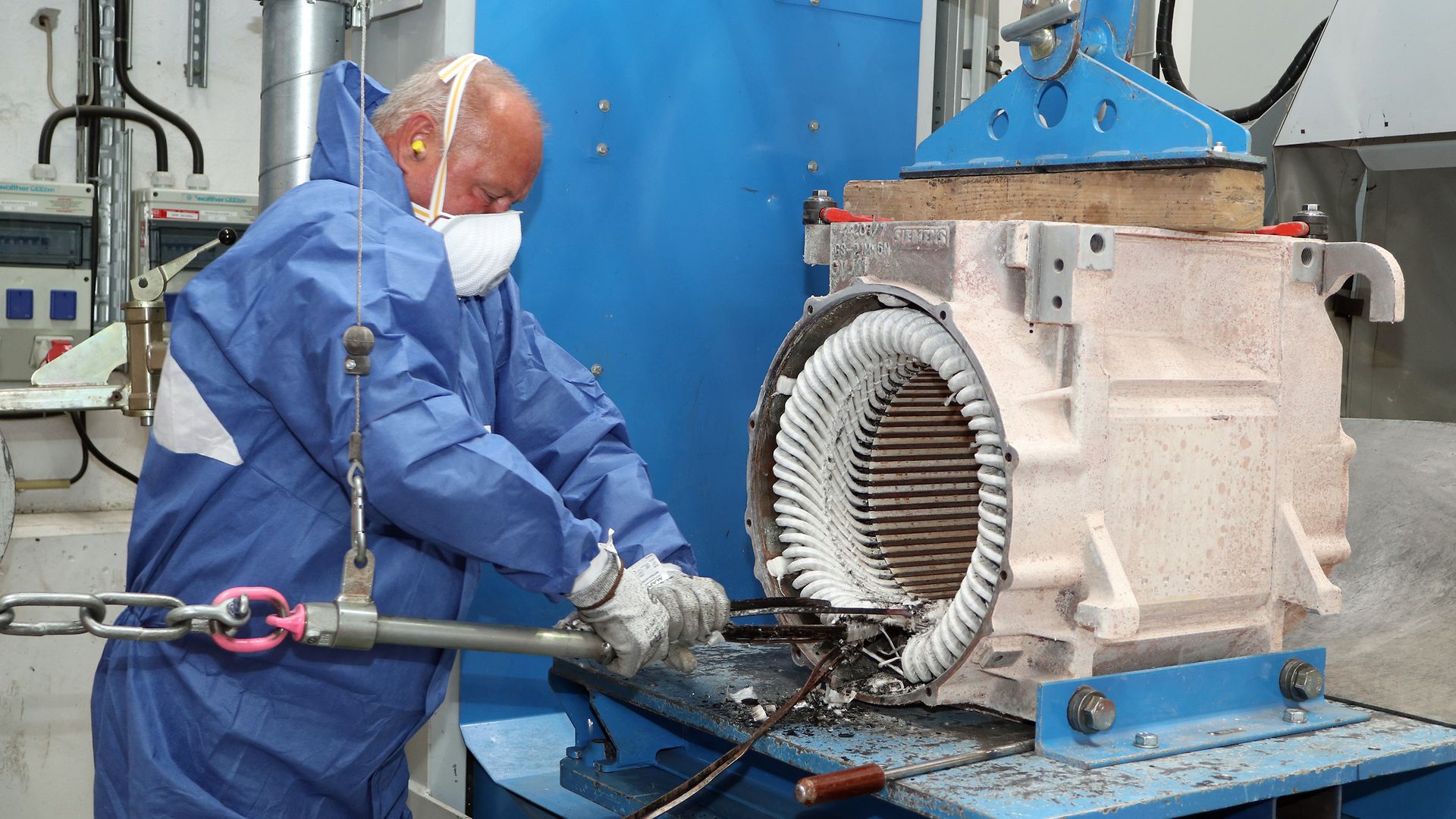 Ein Mitarbeiter in Schutzkleidung und mit Maske entfernt mit einem Werkzeug die alten Wicklungen im Elektromotor.