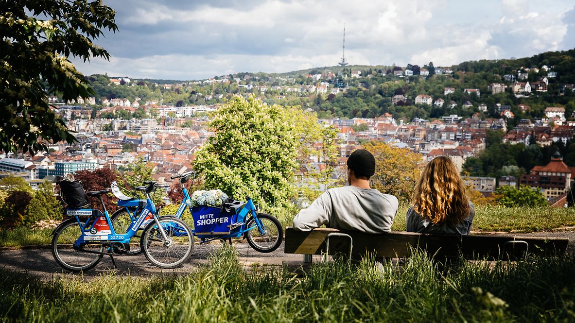 Ein Paar sitzt auf einer Bank und schaut ins Tal hinab auf die Stuttgarter Innenstadt. Neben der Bank stehen Leihräder.