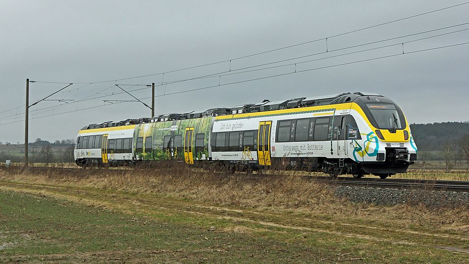 Ein Akkuzug fährt auf Schienen durch eine grüne Landschaft.