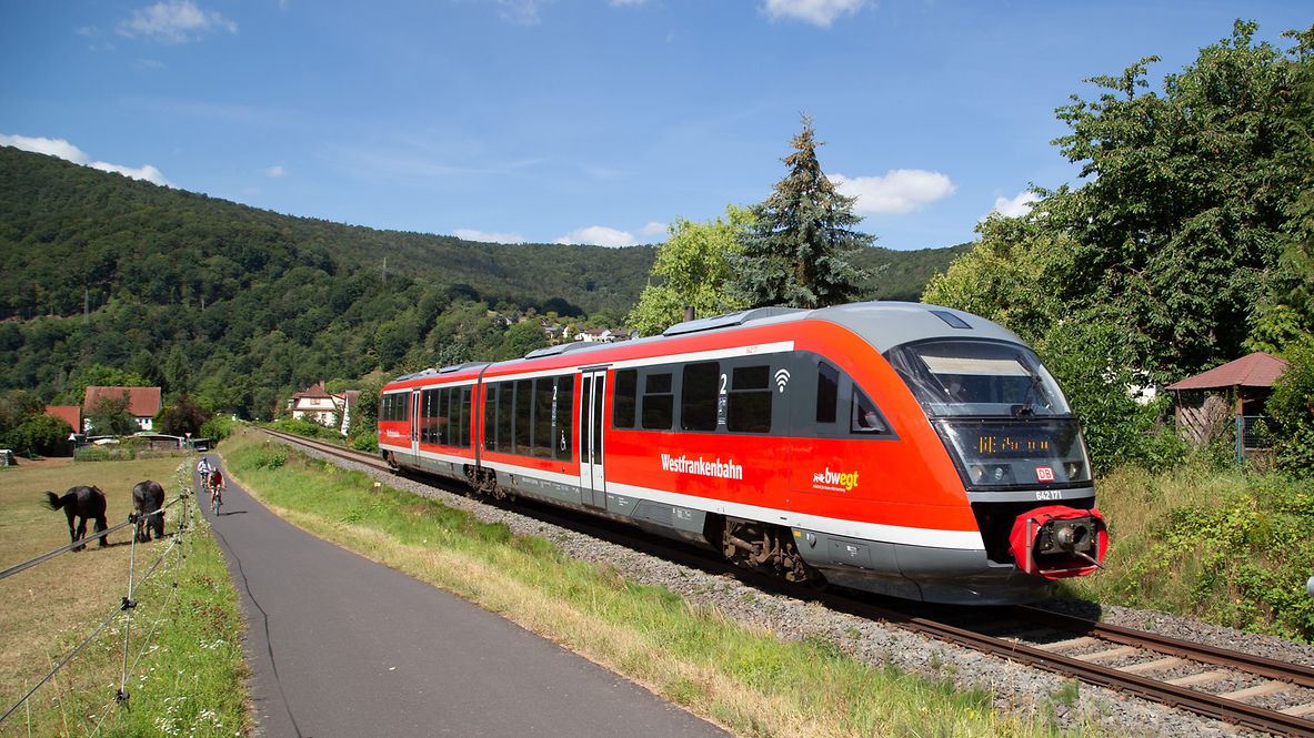 Ein Zug der Westfrankenbahn fährt an zwei Fahrradfahrenden vorbei.