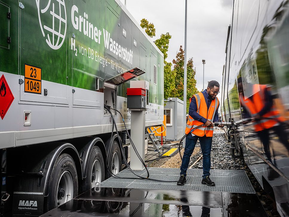 Ein Mann mit Warnweste steht zwischen einem Tanktrailer und Zug und hält einen Tankschlauch in der Hand.