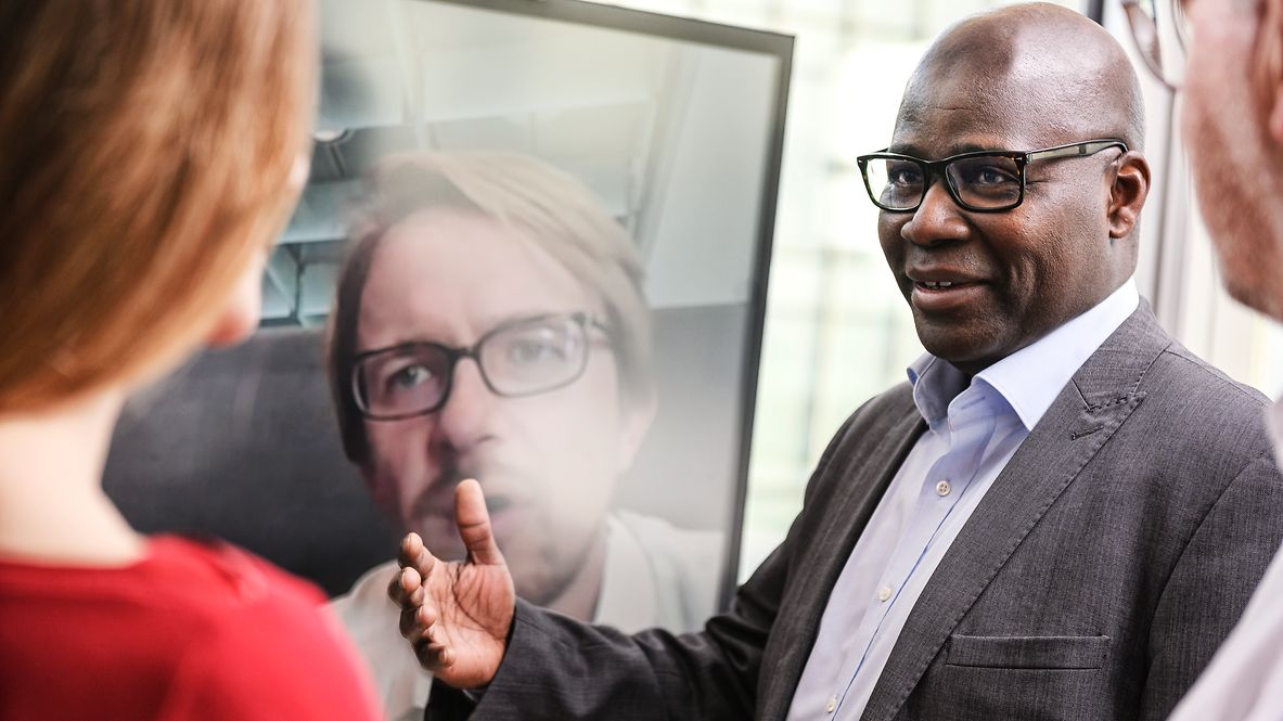 Drei Personen bei einer Besprechung vor einem Monitor. Eine vierte Person ist per Video dazugeschaltet.