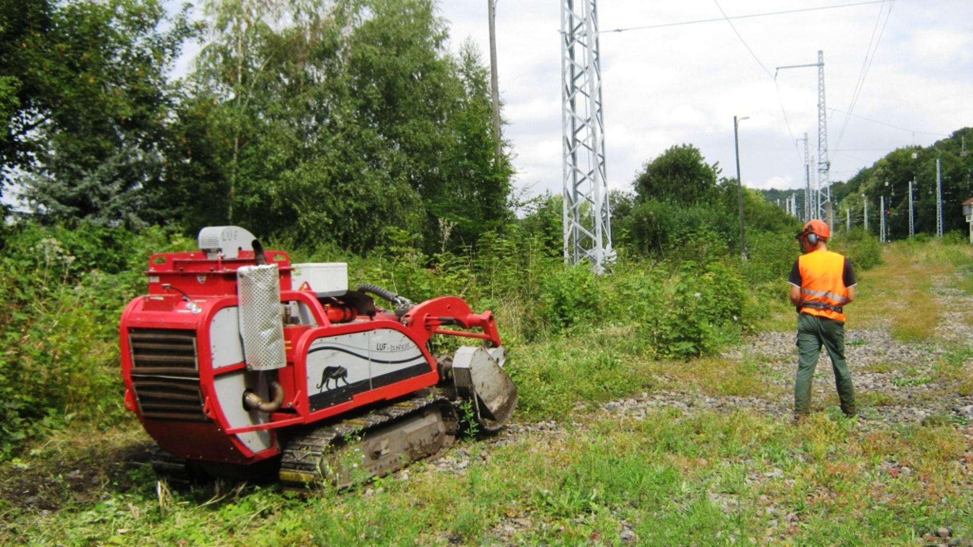 Eine Mulchraupe schneidet die Grünflächen neben einem Gleis frei und wird von einem Bauarbeiter ferngesteuert.