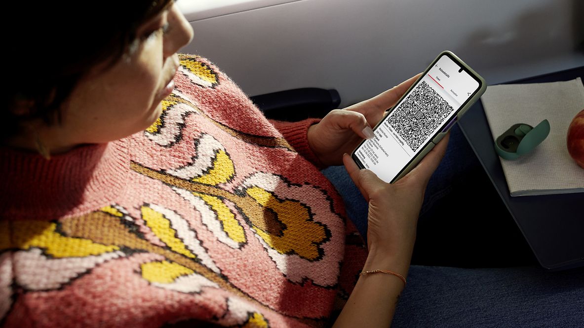 Eine Frau sitzt im Zug und hält ihr Smartphone in der Hand. Auf dem Bildschirm ist der QR Code eines Handytickets zu sehen.