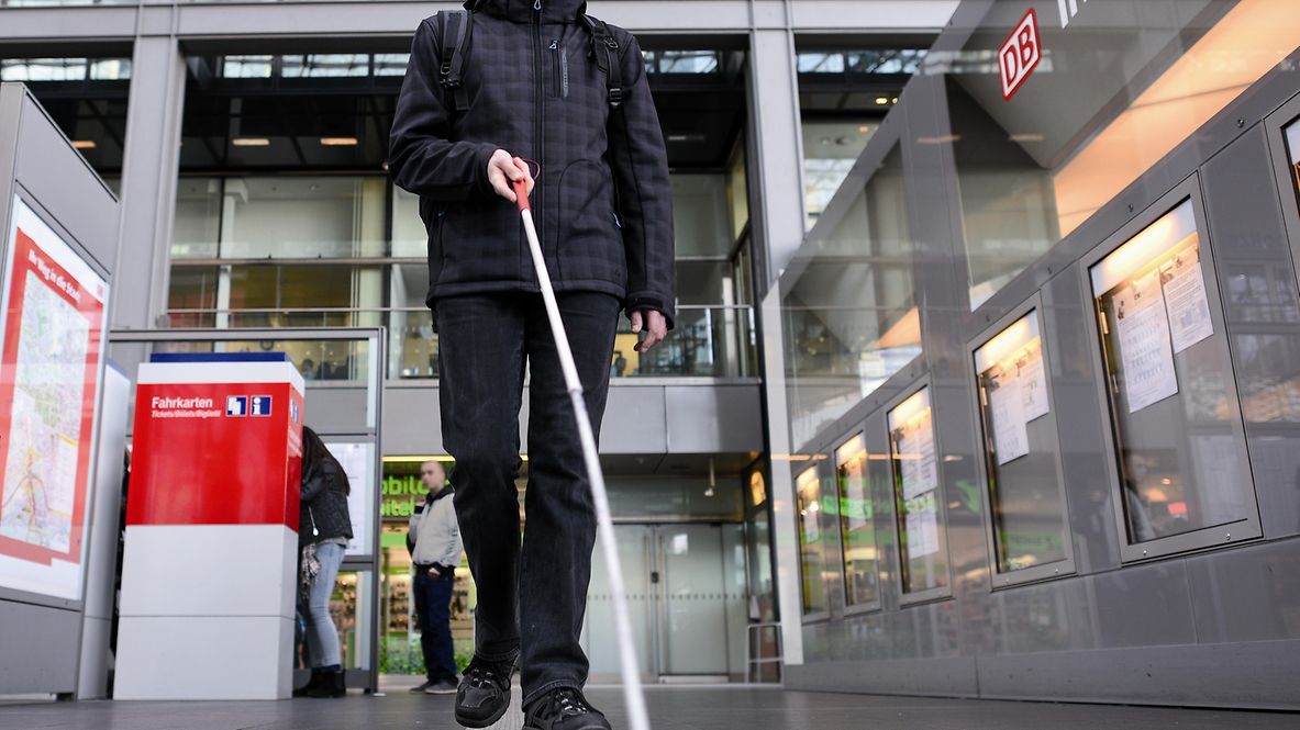 Mann mit Blindenstock orientiert sich an einem taktiles Leitsystem in einem Bahnhof.