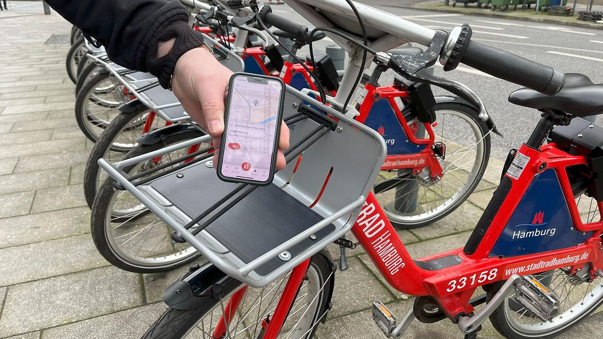 Eine Person zeigt die StadtRAD Hamburg App auf ihrem Smartphone. Im Hintergrund stehen StadtRAD Hamburg Fahrräder.