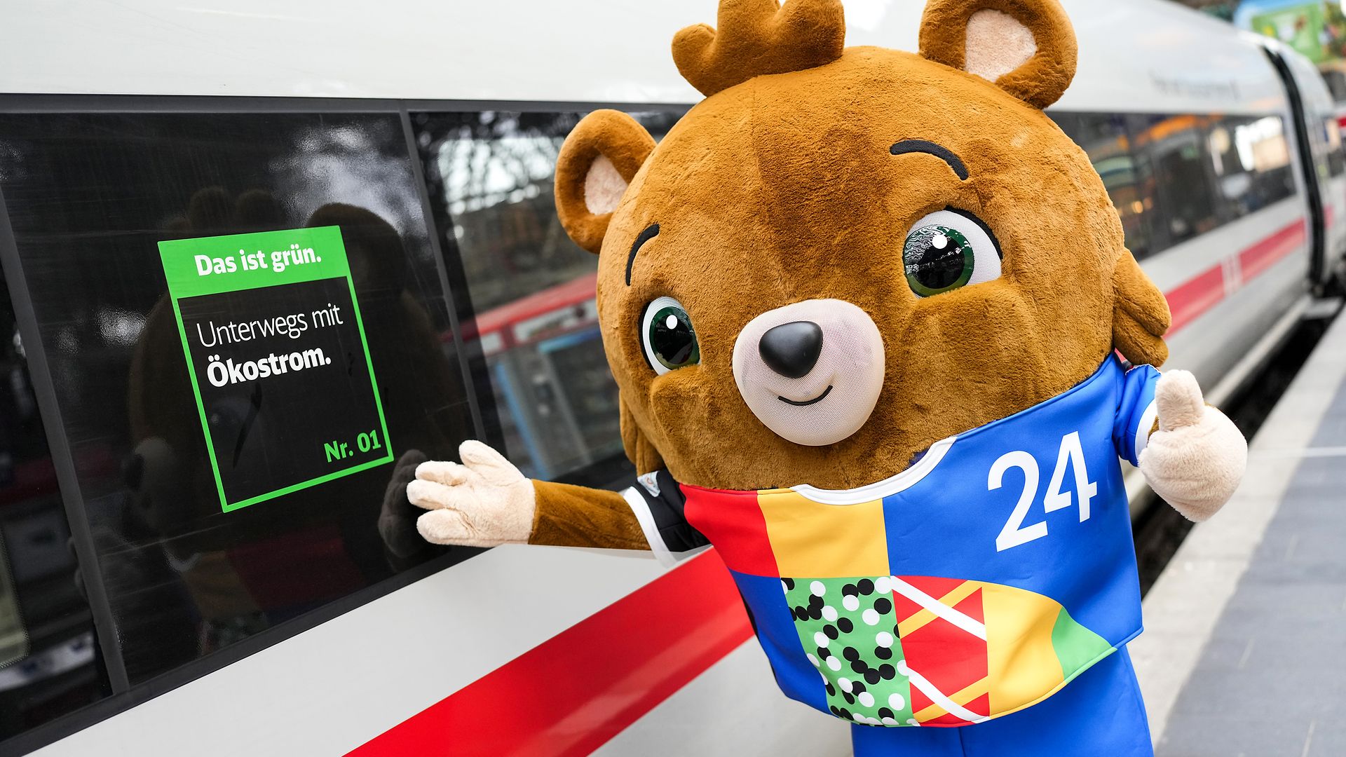 Das Maskottchen für die UEFA EURO 2024™ steht an einem Gleis an einem Bahnhof neben einem ICE.