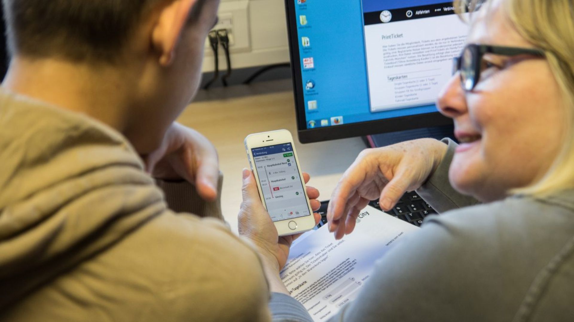 Ein junger Mann und eine ältere Frau beugen sich an einem Schreibtisch gemeinsam über ein Smartphone.