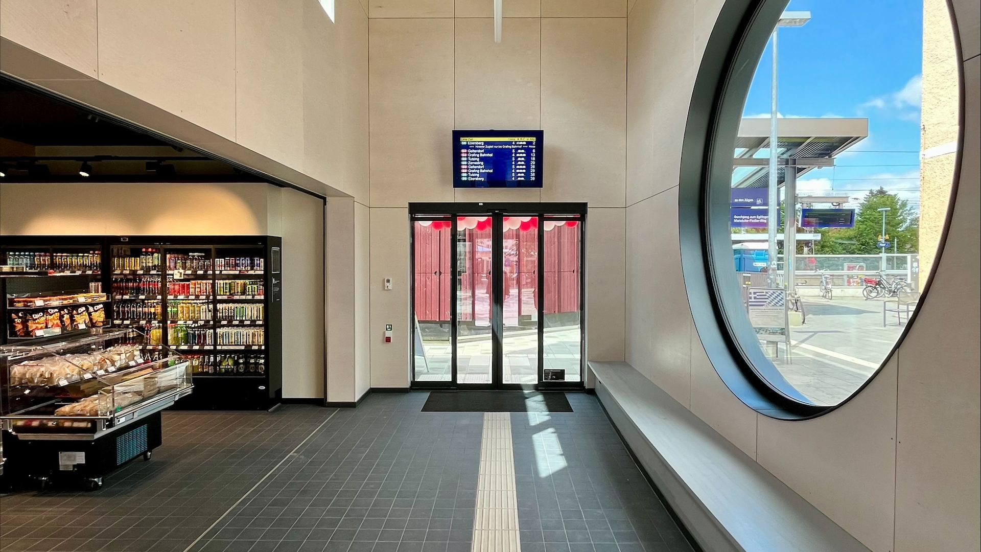 Blick in den Empfangsbereich des Bahnhofsgebäude Haar mit Verkaufsregal, Sitzbank, großem Rundfenster, Glastür und Zuganzeiger.