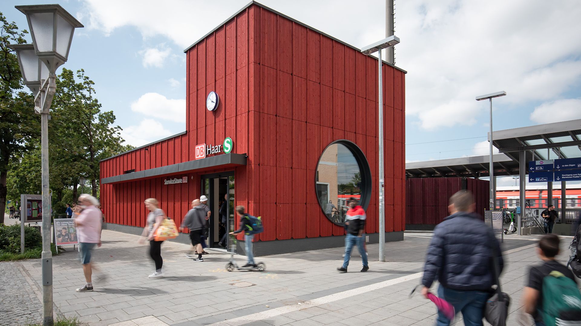 Der Bahnhofsvorplatz in Haar mit rotem Empfangsgebäude im Hintergrund und vorbeilaufenden Passanten im Vordergrund.