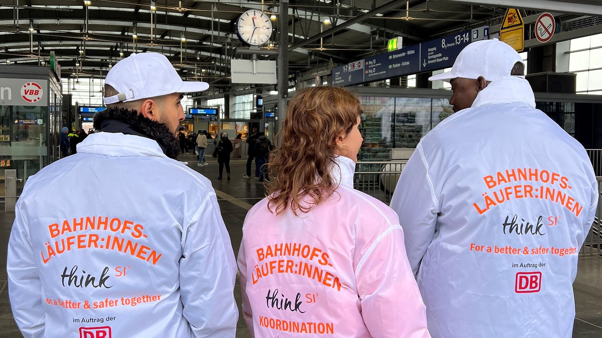 Drei junge Menschen laufen in Jacken, auf denen Bahnhofsläufer:innen steht, über einen Bahnhof. 