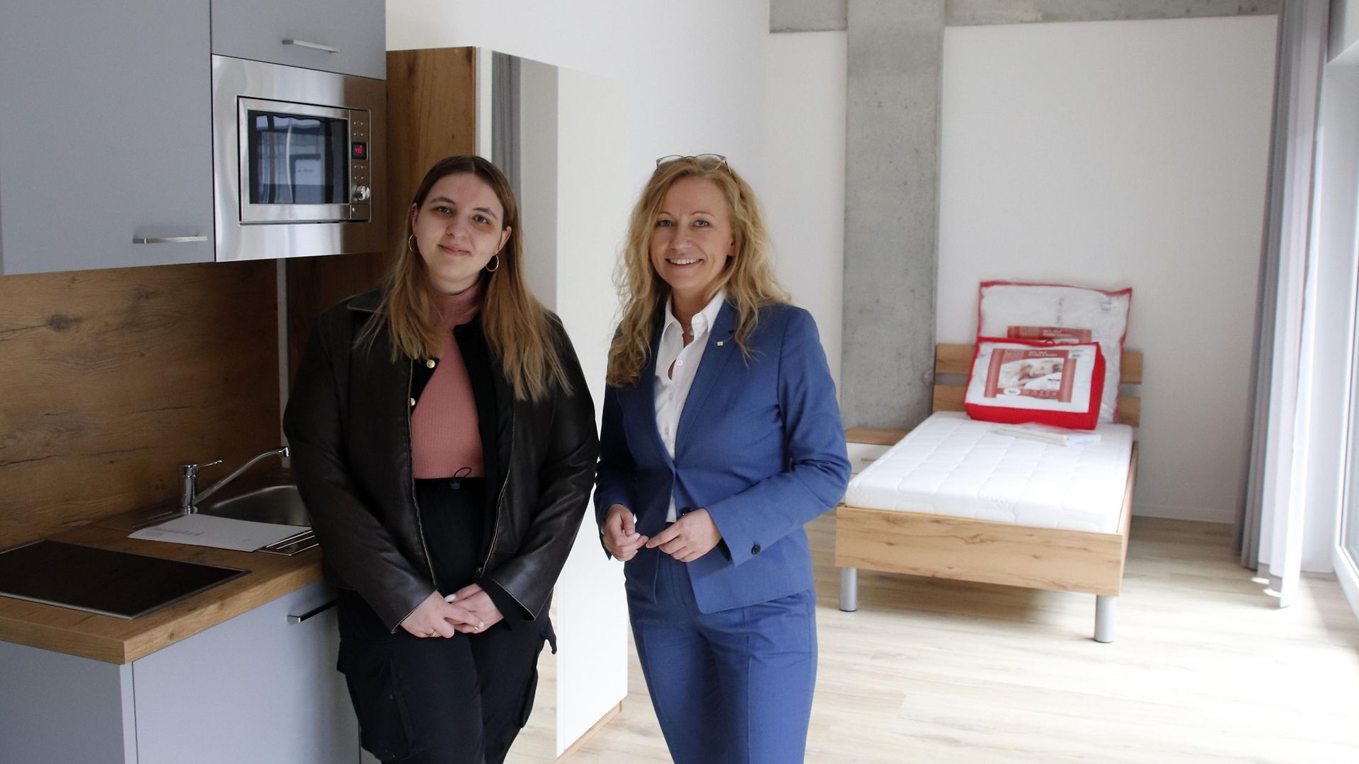 Eine junge Auszubildende und eine mittelalte Frau stehen in einer kleinen möblierten Wohnung.