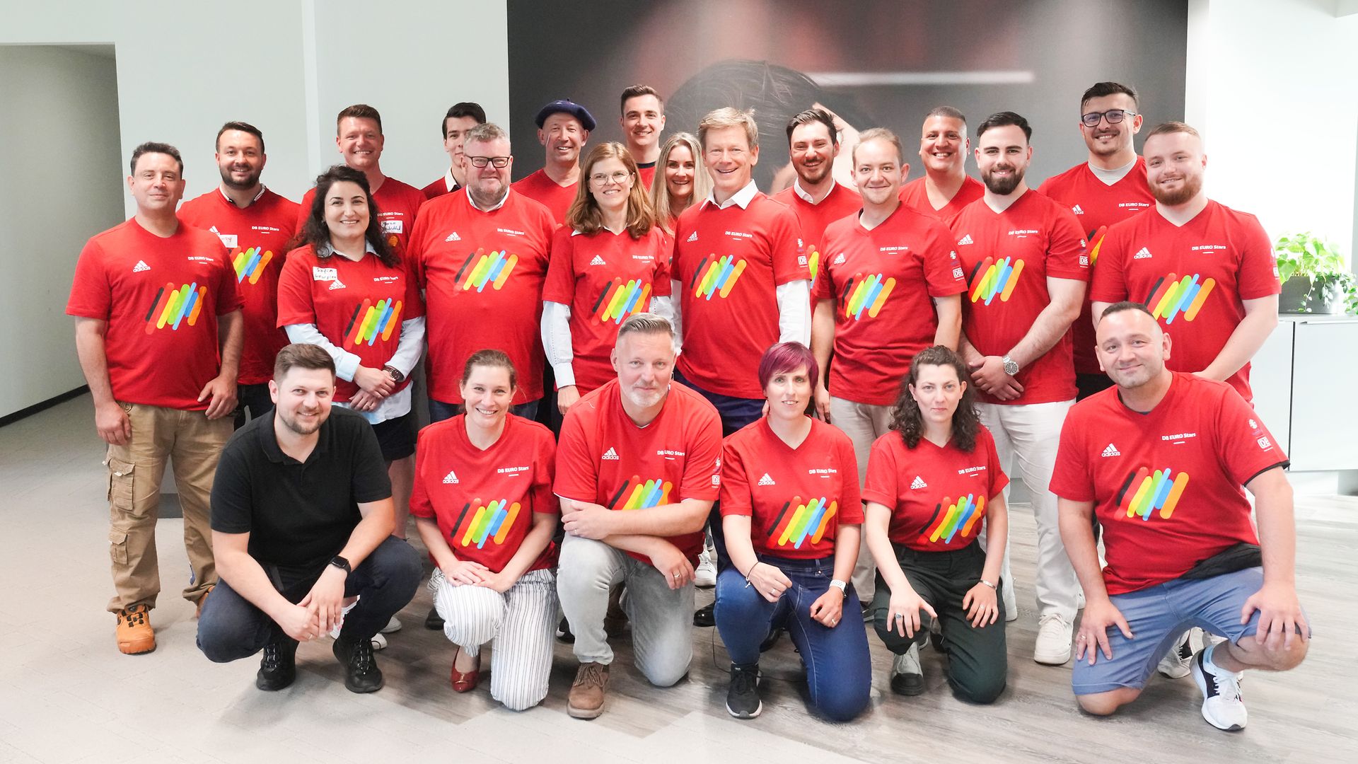 Eine Gruppe von DB-Mitarbeitenden posieren gemeinsam in einem Büro mit roten DB-Trikots.