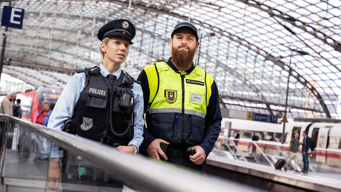Eine Bundespolizistin und ein Mitarbeitender von DB Sicherheit stehen an einem Gleis am Berliner Hauptbahnhof.
