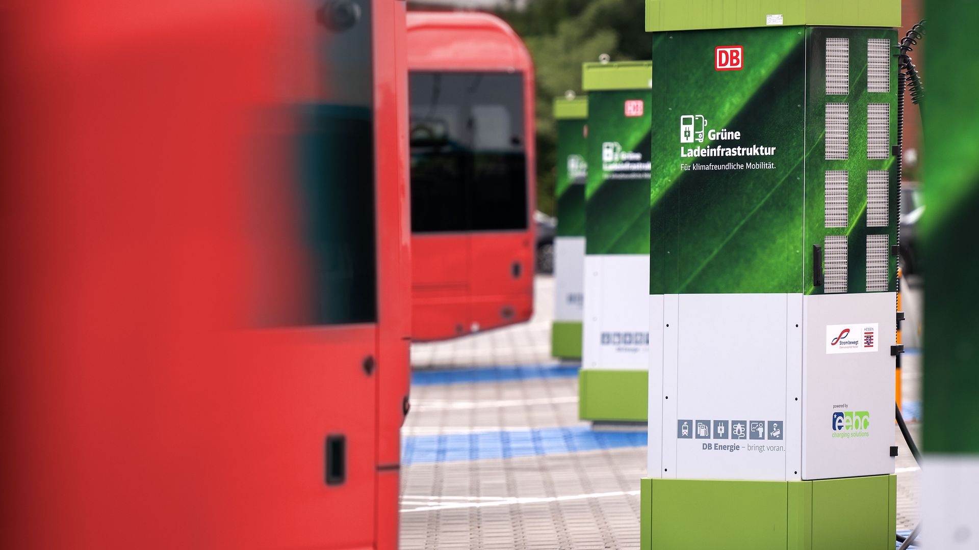 Eine grüne Ladesäule neben roten Elektro-Bussen am Bahnhof 