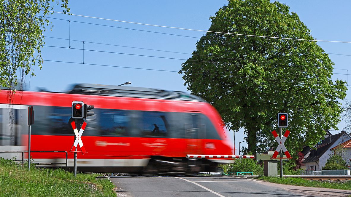 Ein roter Zug fährt im Sommer über einen Bahnübergang, die Schranken sind geschlossen. 
