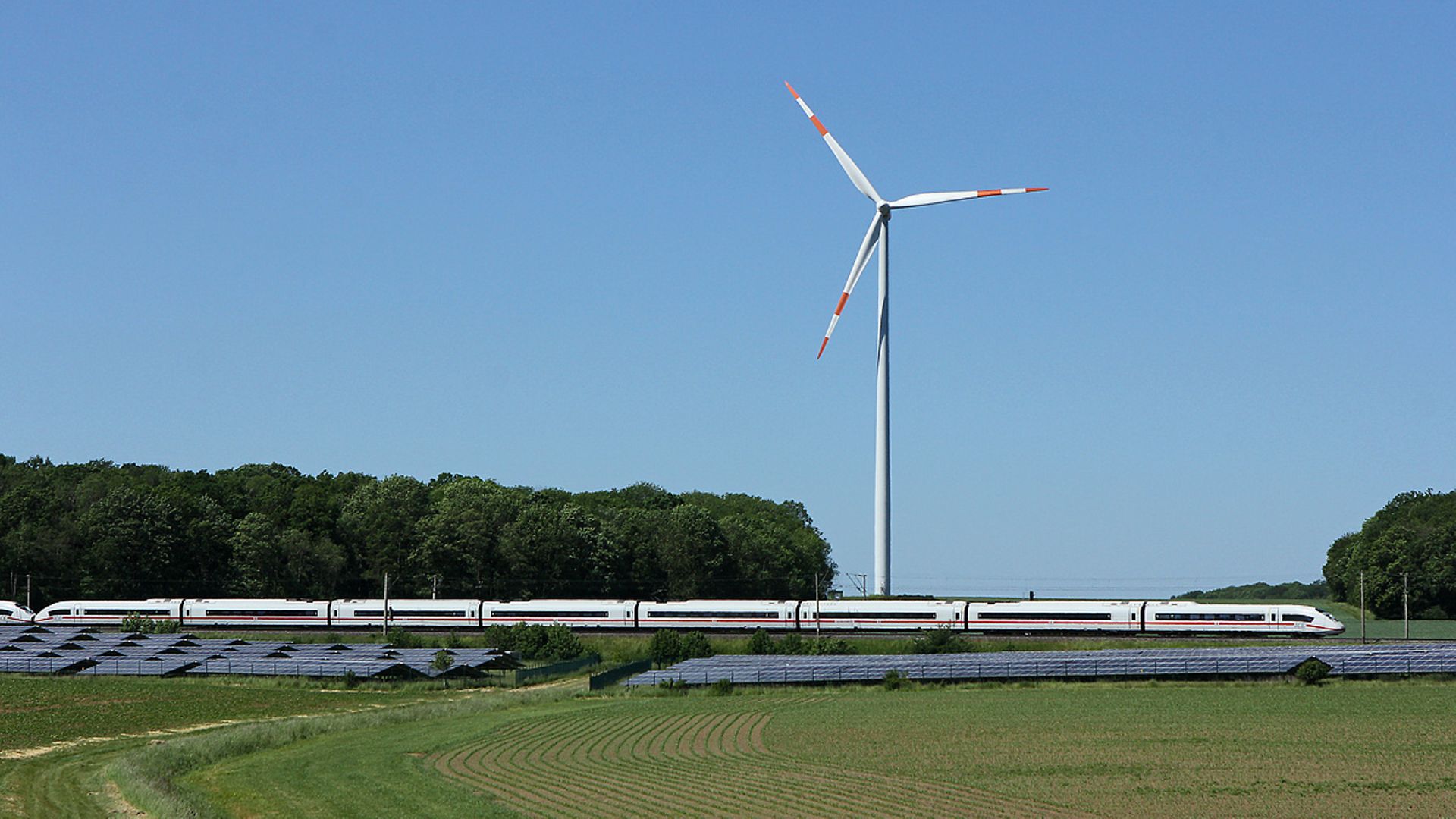 Ein ICE-Zug fährt vorbei an einer Windkraftanlage und einem Solarpark.